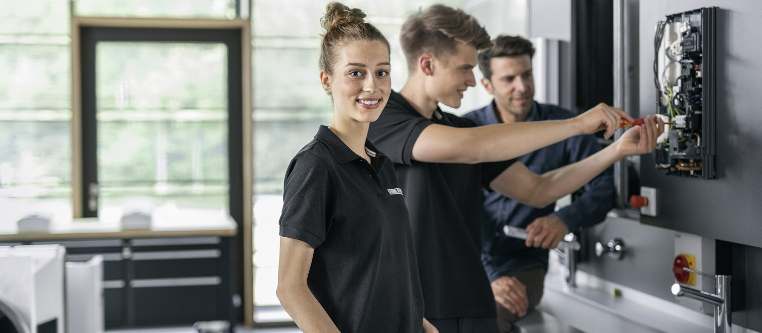 Συμμετέχουσα χαμογελά καθώς τεχνικός εγκαθιστά ταχυθερμαντήρα νερού STIEBEL ELTRON σε εκπαιδευτική δοκιμή