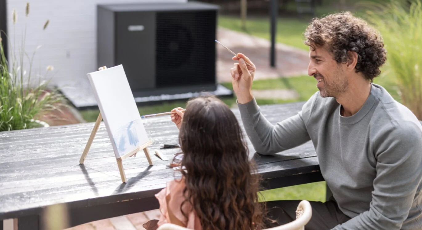 Οικογένεια απολαμβάνει τον εξωτερικό χώρο με διακριτικά τοποθετημένη αντλία θερμότητας STIEBEL ELTRON στο βάθος